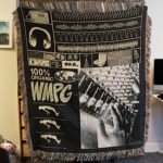 Picture of someone holding up the blanket standing up. The design on the blanket shows different types of sound waves, instrument close up, and other musical imagery.