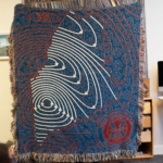 Picture of a blanket being held up, the blanket is mostly blue with red and white lines showing the outline of the state of Maine. On the bottom right side is a WMPG logo in red.
