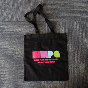 Picture of a black tote bag on a dark carpeted background. On the black tote bag is a blocky WMPG logo in pink, purple, blue, and green.