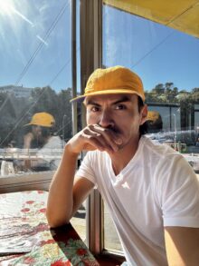 Candid photo of DJ El Perseguidor wearing a yellow cap sitting a table with the sun in corner through a big picture window.